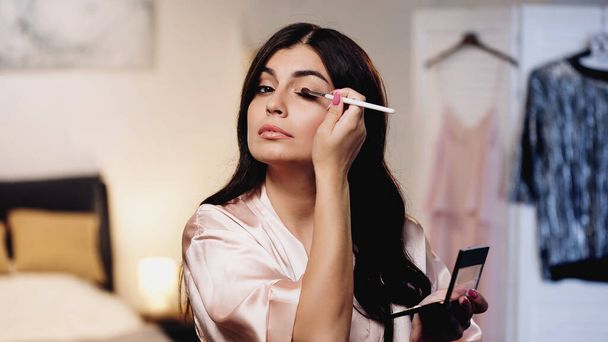 young woman in pink silk peignoir applying eye shadows with cosmetic brush in bedroom - Foto, Imagen
