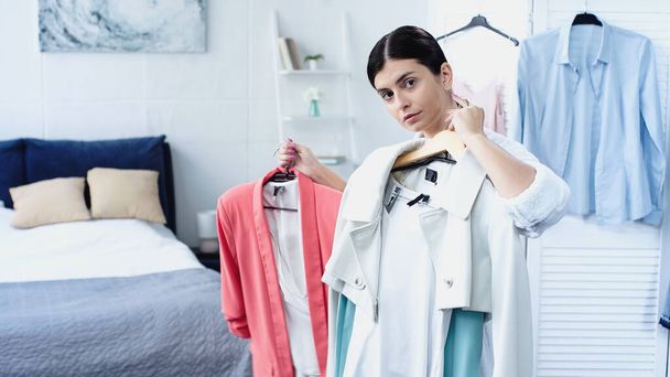 brunette young woman in bathrobe holding clothes on hangers in bedroom  - Photo, Image