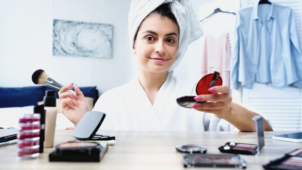 smiling young woman with head wrapped in towel holding cosmetic brush and face powder in bedroom - Photo, Image
