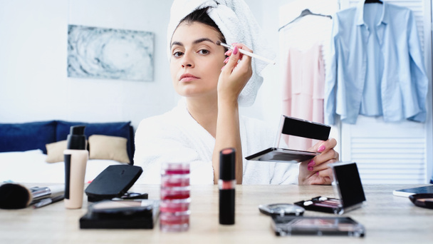 mujer joven en albornoz aplicando sombras de ojos con cepillo cosmético cerca de la mesa con cosméticos decorativos en el dormitorio - Foto, Imagen