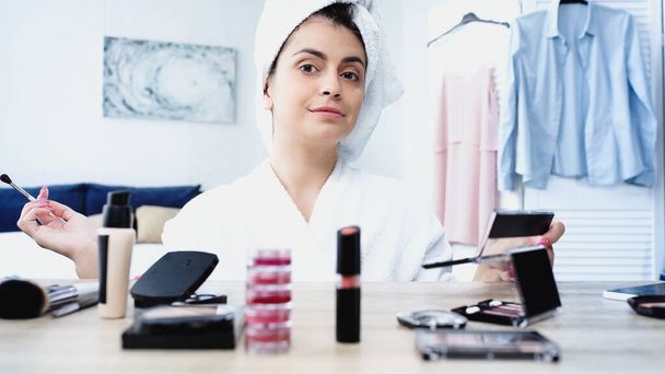 cheerful young woman in bathrobe sitting with cosmetic brush and eye shadows near table with decorative cosmetics in bedroom - Photo, Image