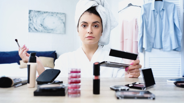 serious young woman in bathrobe sitting with cosmetic brush and eye shadows near table with decorative cosmetics in bedroom - Photo, Image