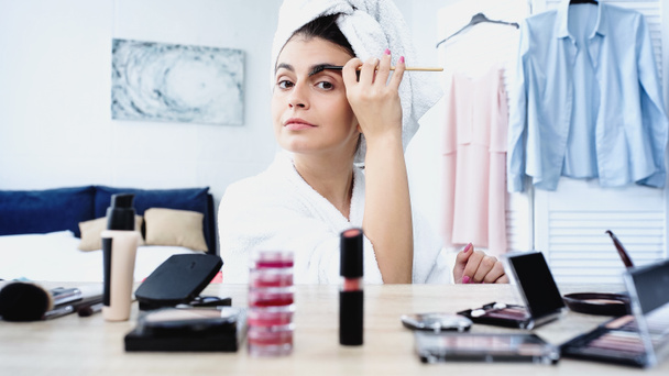 mujer joven en albornoz con cabeza envuelta en toalla estilo ceja con cepillo cerca de la mesa con cosméticos decorativos en el dormitorio - Foto, Imagen