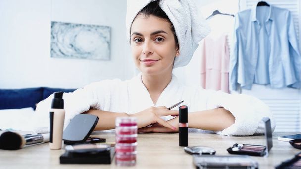 smiling young woman sitting with cosmetic brush near table with decorative cosmetics in bedroom - Photo, Image