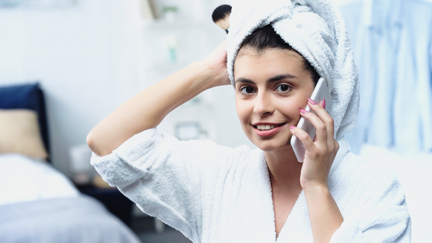 young woman with head wrapped in towel holding cosmetic brush and speaking on cellphone in bedroom - Photo, Image