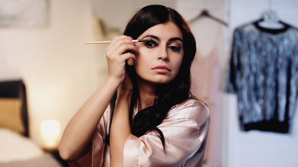 focused young woman in pink silk peignoir styling eyelashes with eyebrow brush in bedroom - Photo, Image