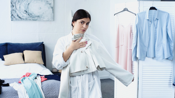 jonge vrouw in witte badjas pasvorm jas in de buurt van bed met kleren in de slaapkamer - Foto, afbeelding