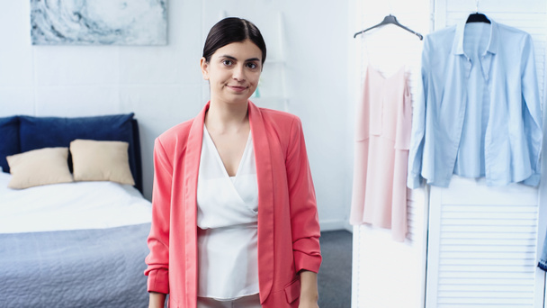 confused young woman standing in white blouse and pink blazer near hangers with clothes in bedroom - Foto, Imagem