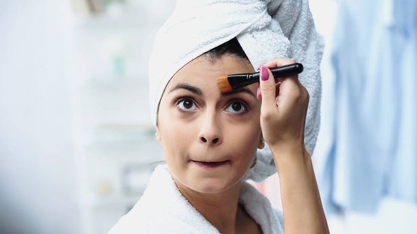 portrait of young woman with head wrapped in towel grimacing and applying foundation on face with cosmetic brush in bedroom - Photo, Image