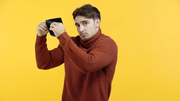 Niezadowolony mężczyzna w swetrze trzyma pusty portfel odizolowany na żółto  - Zdjęcie, obraz