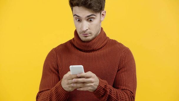 Amazed man in sweater using cellphone isolated on yellow  - 写真・画像