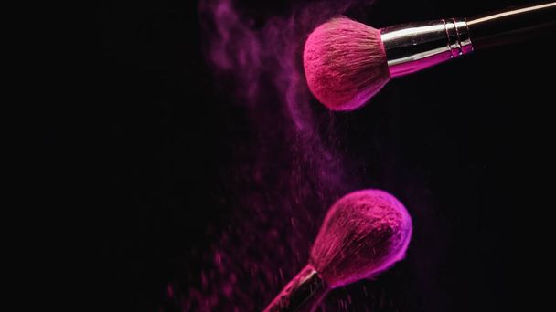 cosmetic brushes with bright pink holi powder near dust splashing on black background - Фото, изображение