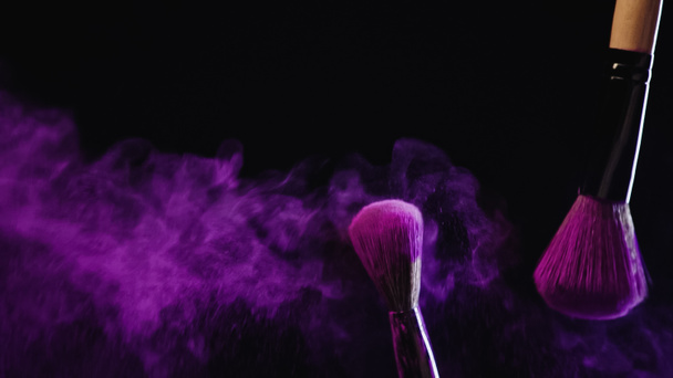 soft cosmetic brushes with purple powder near dust splashing on black background - Foto, Imagen