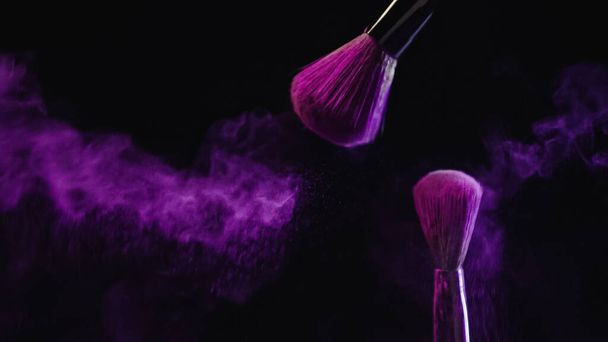 escovas cosméticas com pó roxo vibrante perto de poeira espirrando no fundo preto - Foto, Imagem