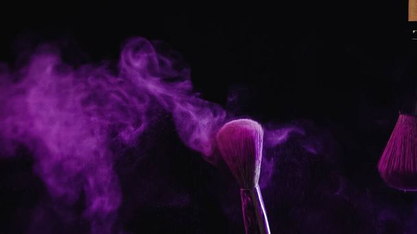 pinceles cosméticos con vibrante pintura holi púrpura cerca del polvo salpicando sobre fondo negro - Foto, imagen