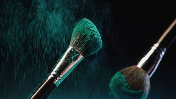 escovas cosméticas com tinta holi turquesa brilhante perto do respingo de poeira no fundo preto - Foto, Imagem