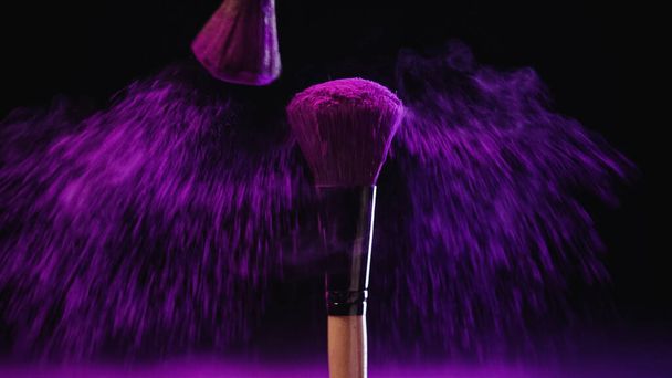 pinceaux cosmétiques lumineux avec peinture holi près de poussière rose sur fond noir - Photo, image