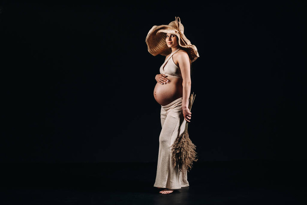 pregnant woman in a straw hat with beige clothes with a bouquet in her hands in the studio on a black background - Fotó, kép