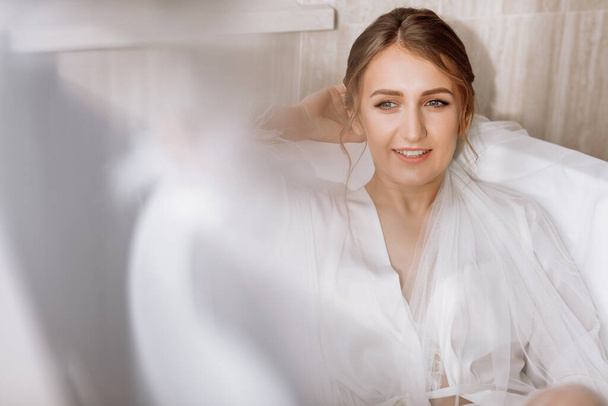 Goedemorgen van de bruid. Gelukkig mooie jonge vrouw draagt in een witte lange sluier, badjas en ondergoed zitten lachen en plezier hebben in het bad op een witte achtergrond, ontspannen. trouwdag. - Foto, afbeelding
