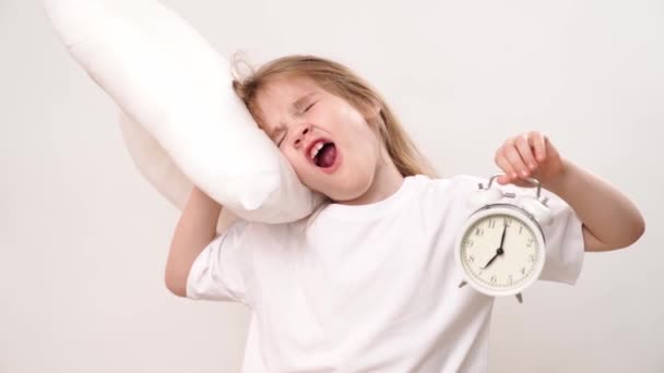小さな眠い女の子が白い背景に枕と目覚まし時計を抱いています。幼児期に学校や幼稚園に通っていました。快適な寝具. - 映像、動画