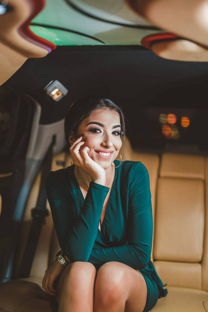 A pretty Caucasian lady with a green evening dress posing while sitting in a luxury limousine - Fotó, kép