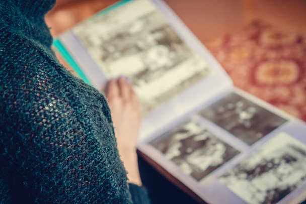 Close-up of a girl in dark clothes sitting on a sofa, holding an album on her lap and looking at old photos. Photo from behind copy space - Photo, image