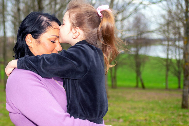 Hermosa madre e hija se besan en la frente abrazos en un parque de verano naturaleza de perfil mirándose el uno al otro. - Foto, imagen