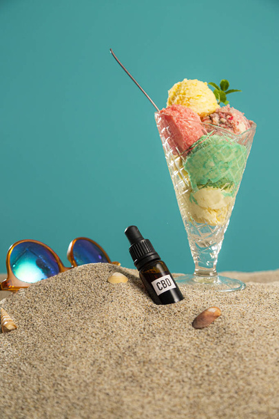 Crème glacée provenant des produits du CBD dans un bol, huile de CBD dans une compte-gouttes et brindilles de la plante de marijuana dans le sable. Le fond est à moitié dans le sable, tandis qu'une partie est quelque chose comme un mur bleu. - Photo, image