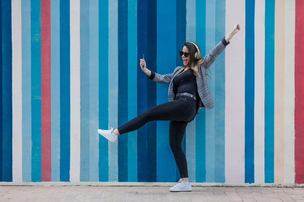 Полностью возбужденное женское тело танцует с поднятой рукой и делает селфи, слушая музыку в наушниках против красочной полосатой стены на городской улице - Фото, изображение