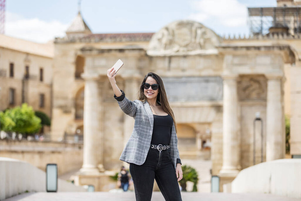 Allegro femmina in abiti casual intelligenti e occhiali da sole sorridente e scattare selfie contro cancello Puerta del Puente durante il viaggio a Cordova, Spagna - Foto, immagini