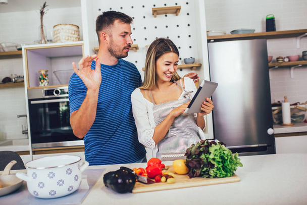Junges glückliches Paar genießt und bereitet gesunde Mahlzeiten in der Küche zu und liest Rezepte auf dem digitalen Tablet. - Foto, Bild