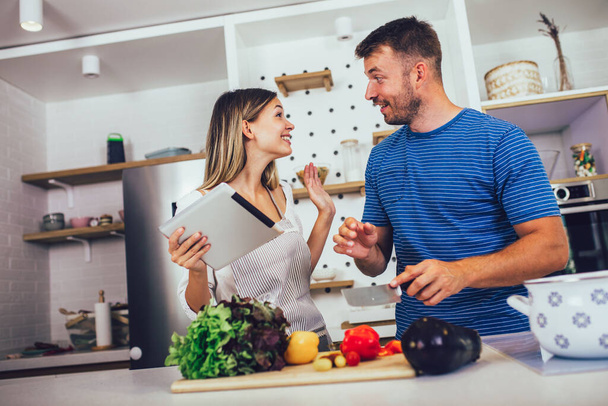 Jovem casal feliz está desfrutando e preparando refeição saudável em sua cozinha e lendo receitas no tablet digital. - Foto, Imagem