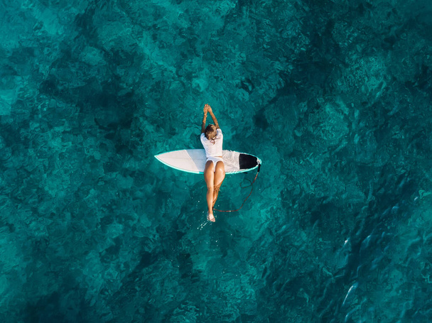 Atractiva chica de surf relajándose en la tabla de surf en el océano transparente. Vista aérea con mujer surfista - Foto, imagen
