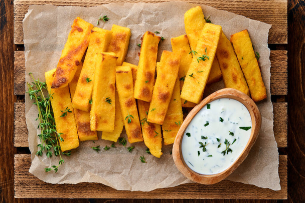 Homemade polenta chips fries with sea salt, parmesan, thyme, rosemary with yogurt sauce. Typical Italian fried polenta. Fried corn sticks. Wooden background. Top view. - Фото, изображение
