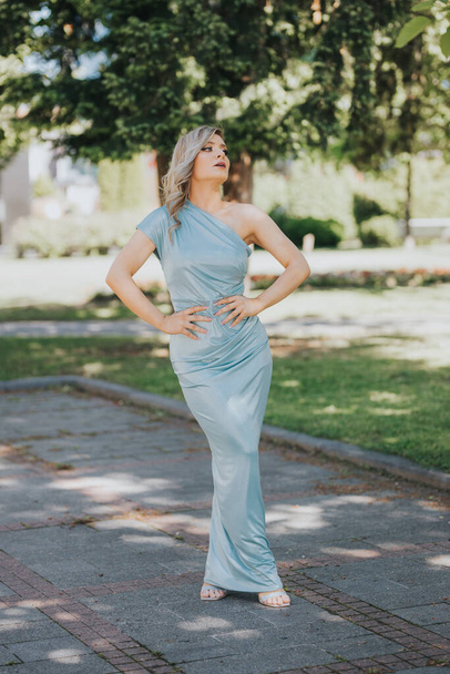 A young and sexy Caucasian female in a long baby blue dress posing in the park - Photo, image
