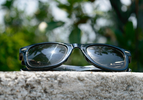 Black sunglasses with summer sunlight reflection placed on wall of a building with lens flare. Close up. Blurry defocused background. UV protection from ultraviolet light and sunburn heat concept. - Fotó, kép