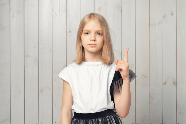 Portret van een schattig blond tienermeisje, wijzend met de vinger omhoog en advies gevend, zegt Aandacht alsjeblieft, luister naar me. Studio shot, lichte houten achtergrond - Foto, afbeelding