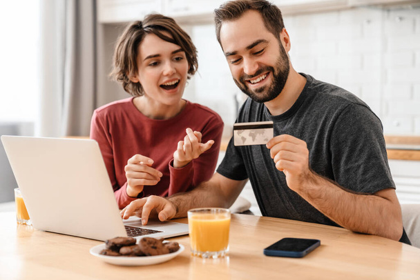 Happy young couple using laptop and credit card while sitting at home - Photo, Image