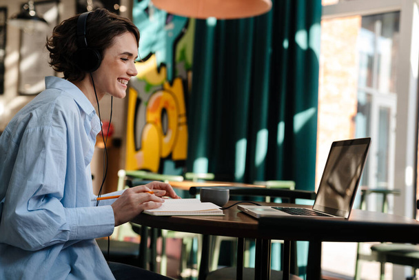 Happy girl wearing headphones making conference call on laptop while sitting in cafe indoors - 写真・画像