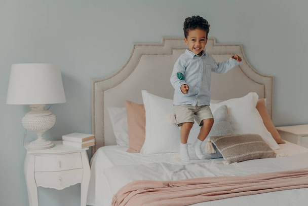Energiczny afro amerykański dzieciak skacze z lizakiem na materacu łóżkowym - Zdjęcie, obraz