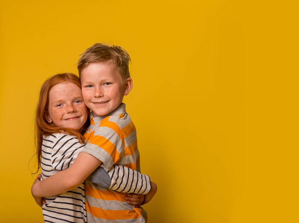Щасливі діти-імбир дівчата і хлопчик обіймаються ізольовано на жовтому тлі. Дитина в смугастій сорочці. Брат і сестра посміхаються і приймають. Літній табір для дітей
. - Фото, зображення