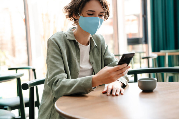 Happy girl in protective mask using cellphone while sitting at cafe indoors - Foto, Imagen