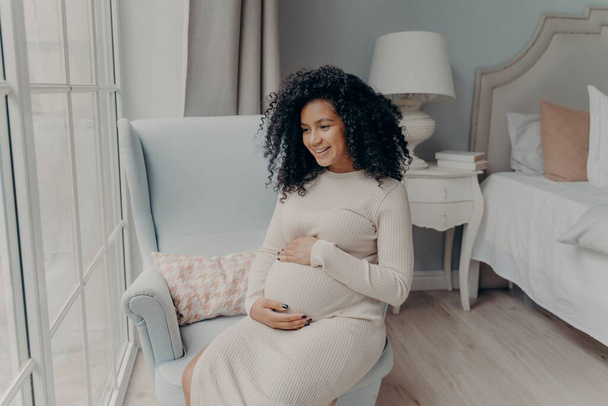 Charmante femme afro-américaine enceinte regardant par la fenêtre et pensant à un futur bébé - Photo, image