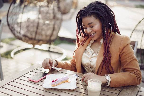 Smiling african american woman drinking coffee and eating dessert in outdoor cafe - Foto, afbeelding