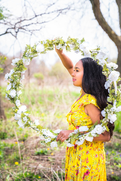 Retrato creativo de una hermosa mujer afroamericana de raza mixta con un vestido amarillo sosteniendo un aro de hula envuelto floral casero enmarcándose dentro y mirando hacia un lado en un soleado día de primavera. - Foto, imagen