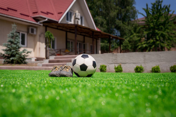 Футбольный мяч и обувь на лужайке на фоне дома с террасой. Главная футбольная концепция - Фото, изображение