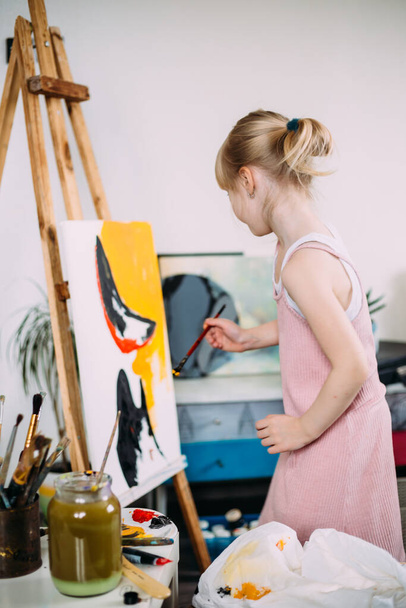Ένα μικρό χαριτωμένο κορίτσι ζωγραφίζει μια μεγάλη εικόνα με ακρυλικό στο σπίτι σε ένα καβαλέτο.  - Φωτογραφία, εικόνα