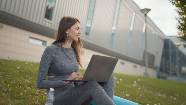 Femme attrayant occupé travaillant à l'ordinateur portable comme assis sur l'herbe dans le parc de la ville. Étudiant étudiant en plein air. Freelance travaillant dans le parc. Fille de fitness athlétique pendant l'entraînement de travail à distance - Photo, image