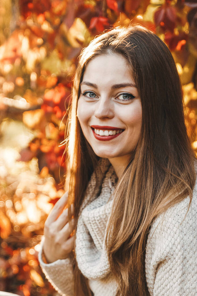 Ritratto ravvicinato di una donna sullo sfondo di foglie rosse e gialle nel parco. Sta indossando un maglione beige e ha un rossetto rosso. Lei sorride e i suoi capelli sono lunghi. - Foto, immagini