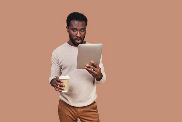 Komea nuori afrikkalainen mies rento vaatteet käyttäen digitaalinen tabletti ja tilalla kuppi kahvia - Valokuva, kuva